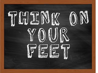 Image of handwritten white chalk text saying "Think on Your Feet" in block capitals with 3d effect on black chalkboard