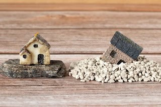 Image of two miniature houses: one sitting upright on a stone slab, the other tilting over on a bed of pebbles